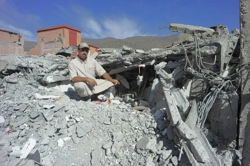 the aftermath of an earthquake with a man sitting on the remains of his house