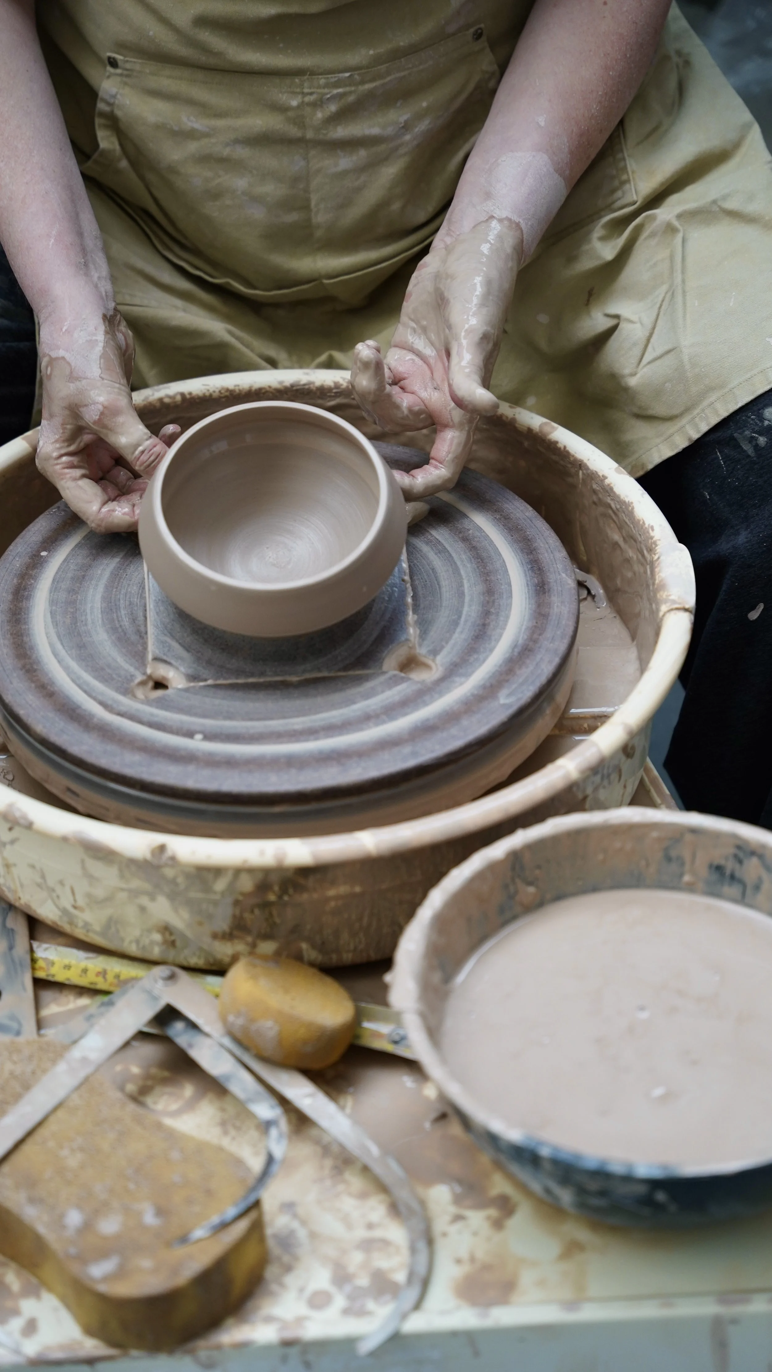 Person shaping a clay cup on a pottery wheel in a pottery studio. The workspace has clay and pottery tools.