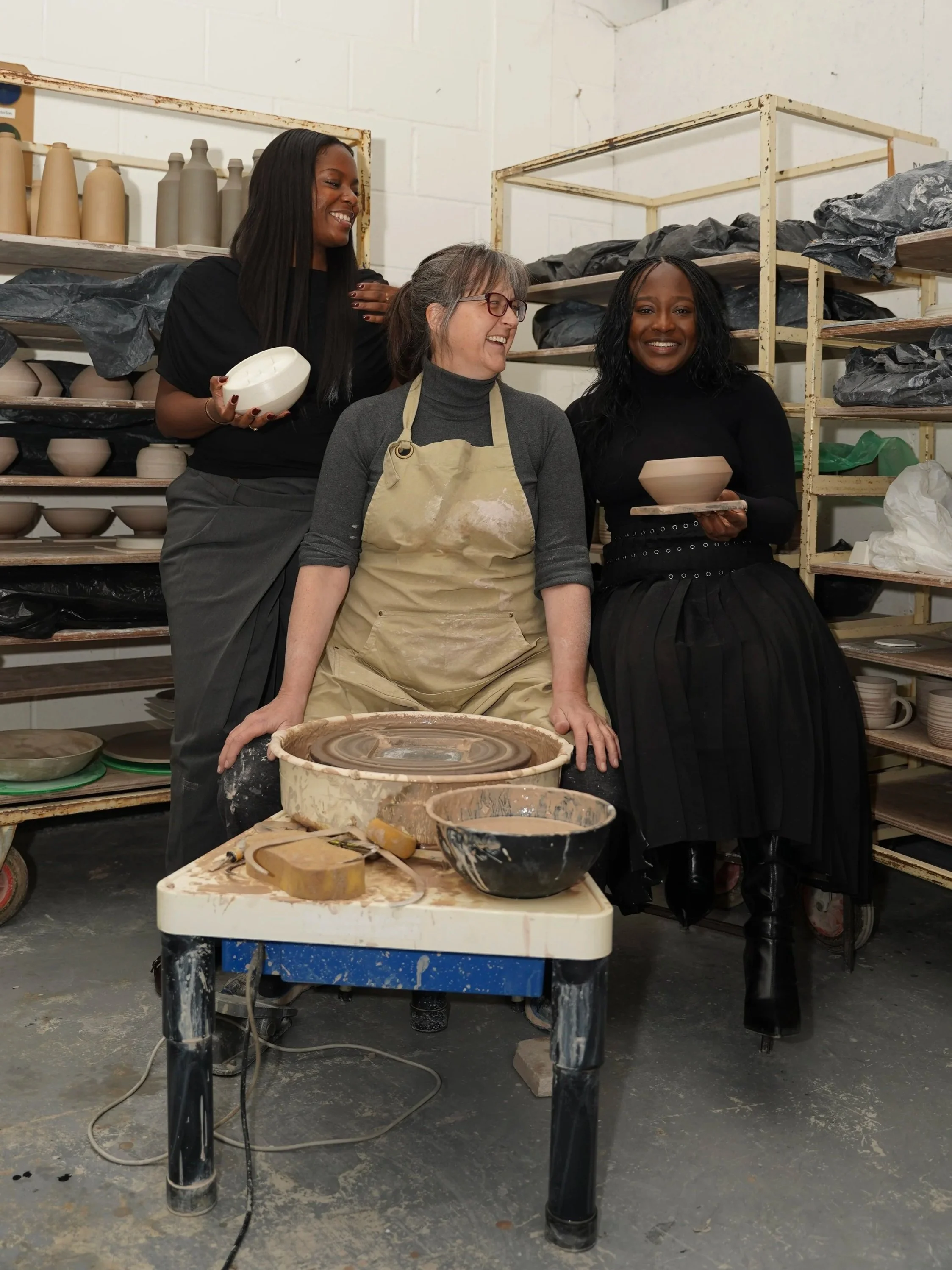 Three women in a ceramics studio, one seated at a pottery wheel, two standing behind her, all smiling and holding pottery pieces.