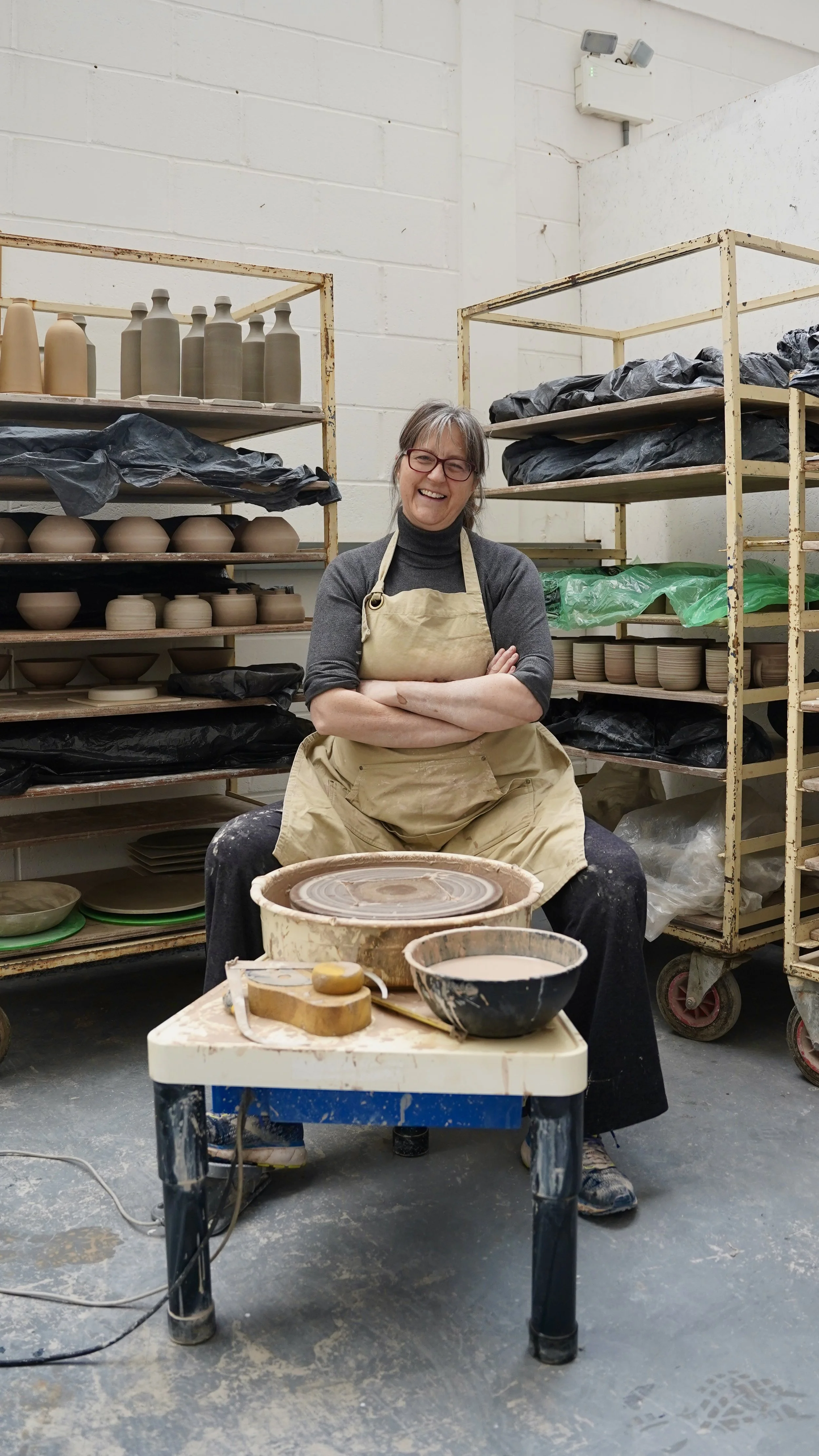 Woman smiling, sitting cross-legged in a pottery studio with shelves of ceramic pieces behind her, a pottery wheel in front, and various pottery tools and bowls on a table.