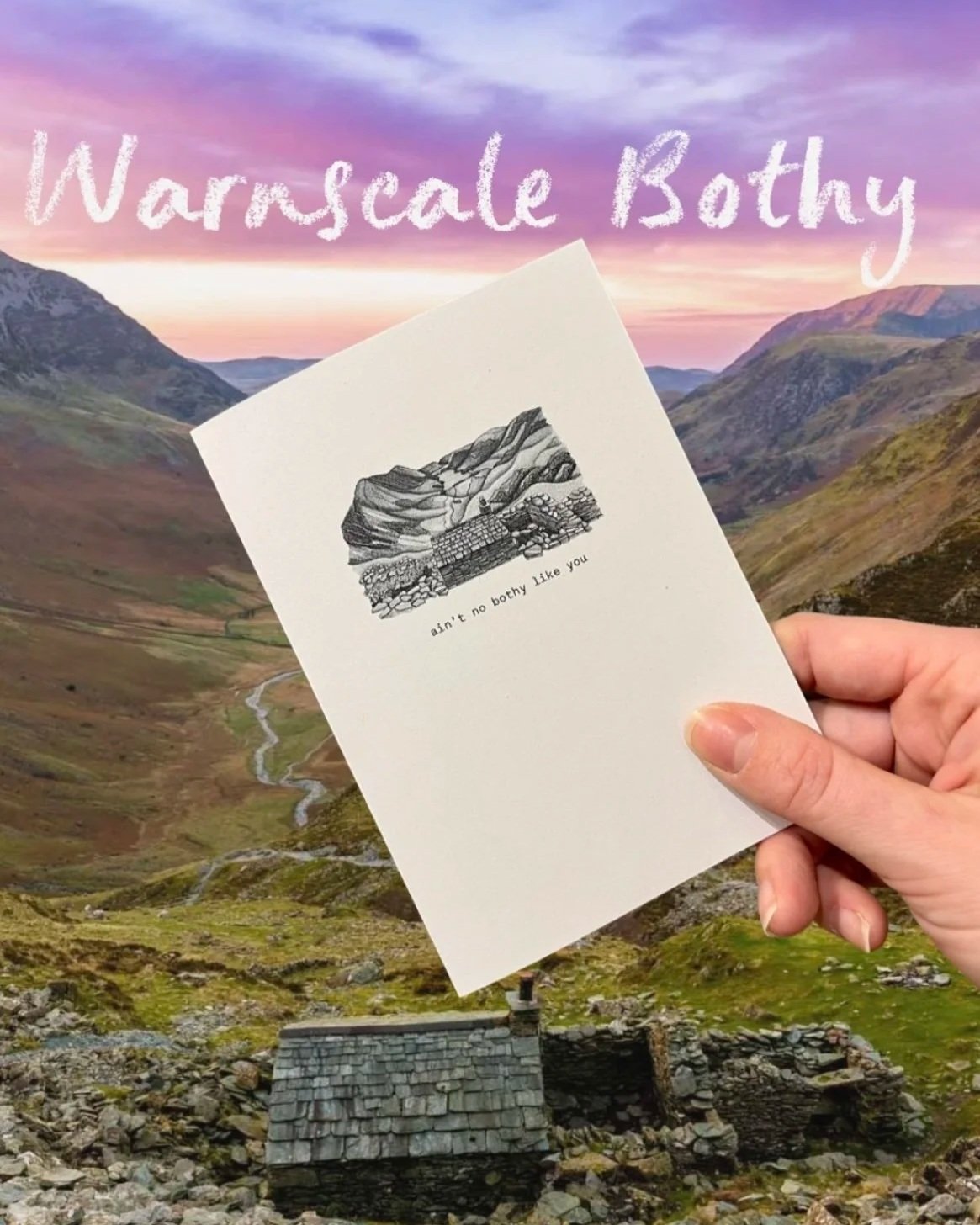 A person holding a card with a black and white illustration of mountains and a stone structure, with text that reads 'ain't no bothy like you', standing in a mountainous landscape at sunset.