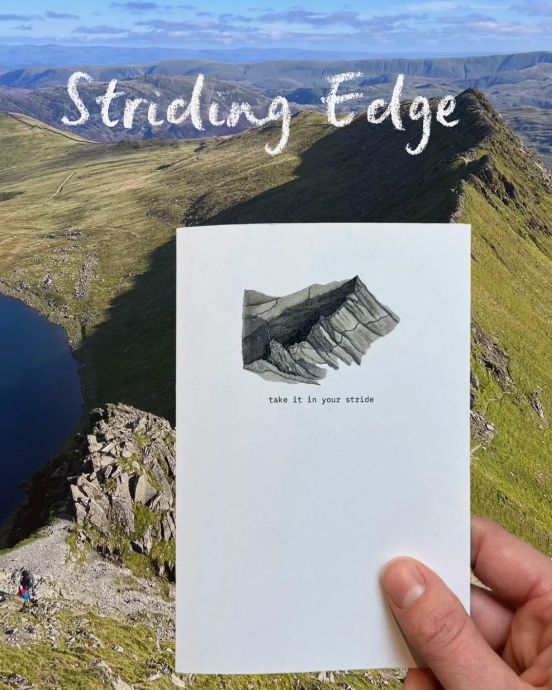 A person holding a booklet labeled "Striding Edge" with an illustration of a mountain ridge and the phrase "take it in your stride." Behind, there is a rugged mountain landscape with steep green slopes, rocky terrain, a small lake, and a trail with h
