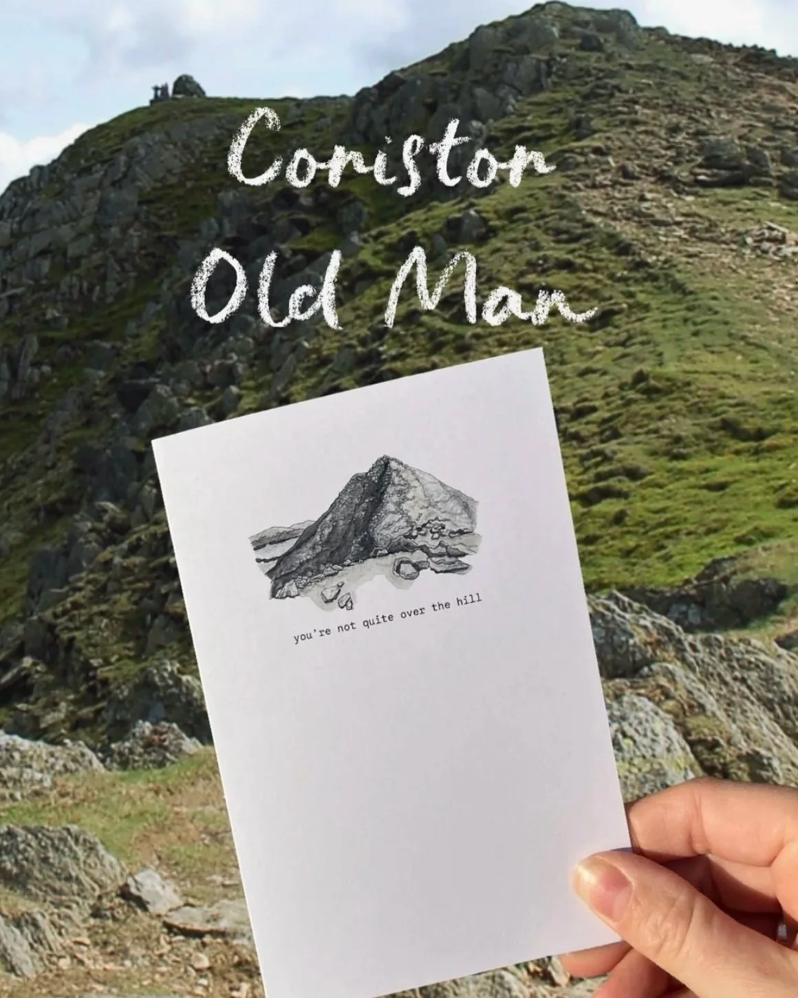 Hand holding a postcard with a black and white sketch of a hill and the text 'you're not quite over the hill' at the base, against a backdrop of a grassy, rocky hillside.