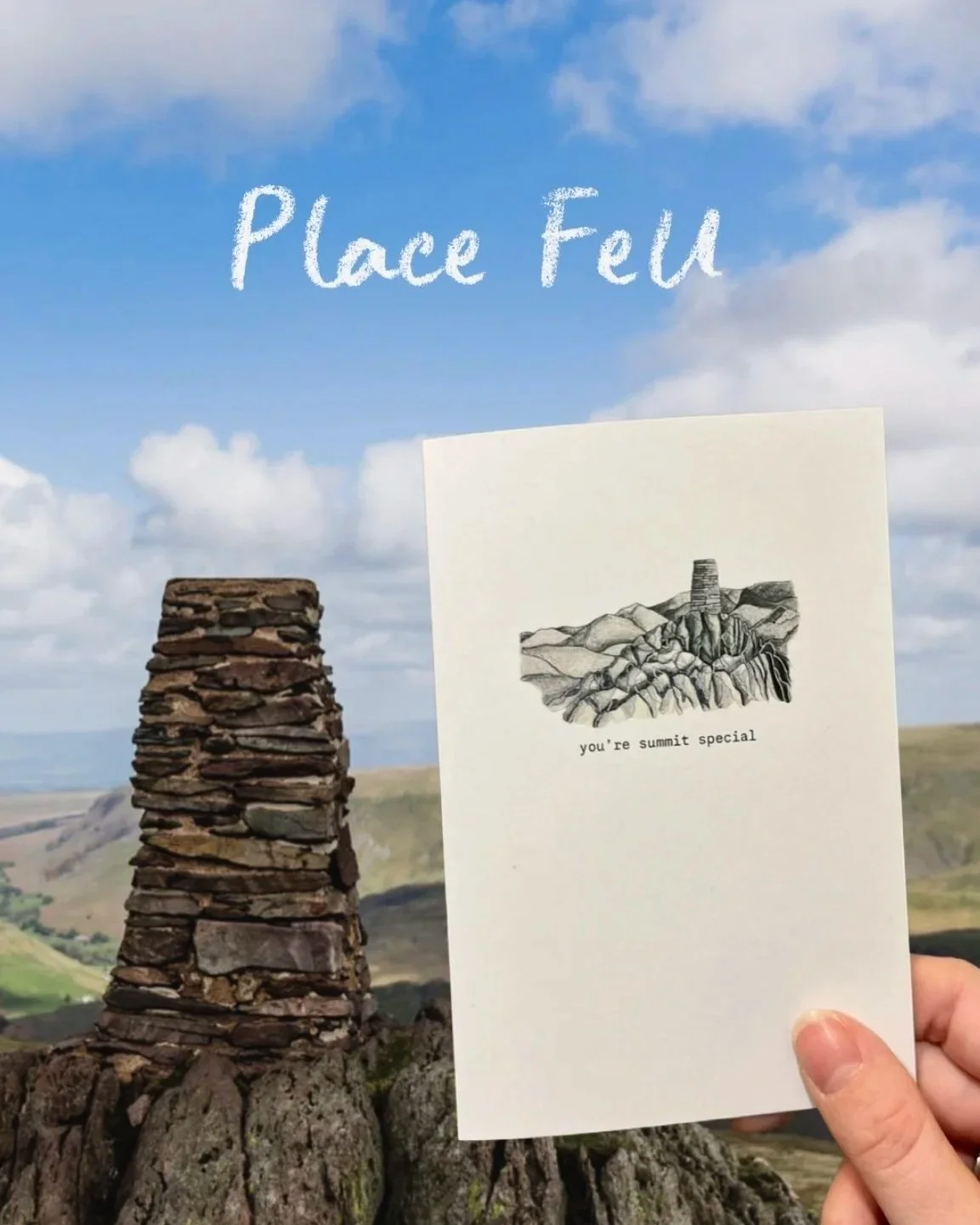 A hand holding a greeting card with a line drawing of a mountain landscape and the phrase "you're summit special" in front of a stone cairn and a scenic hillside landscape under a blue sky with clouds.