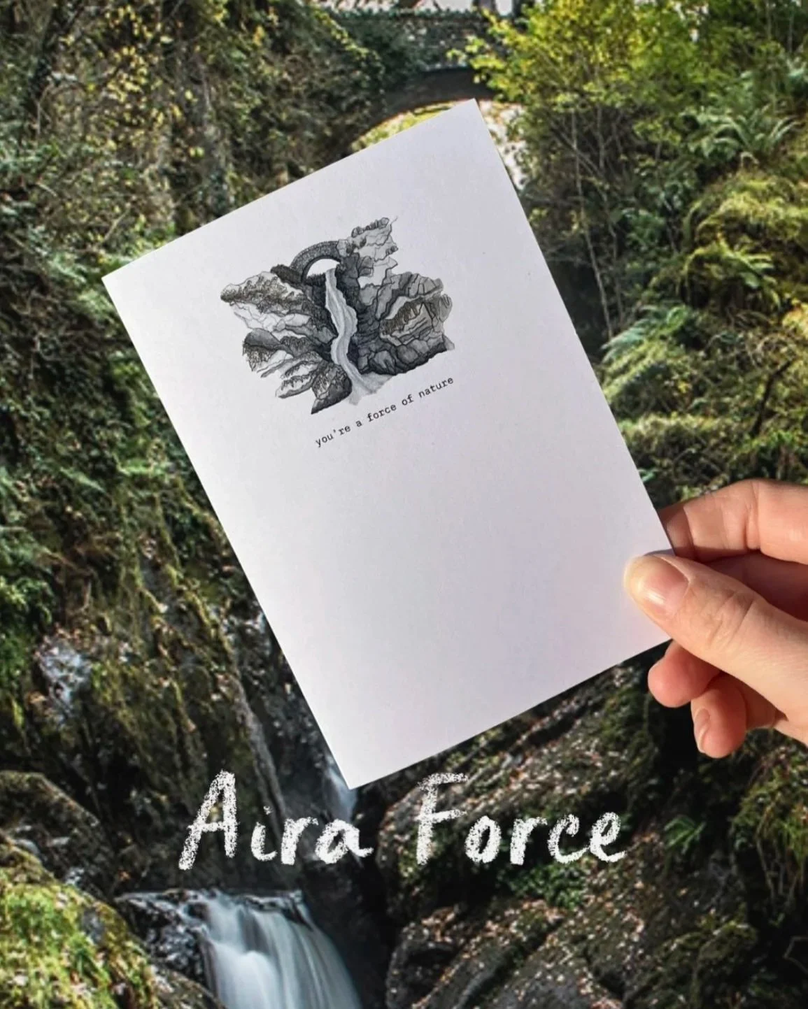 Hand holding a white card with a black and white illustration of a waterfall and trees, along with the text "you're a force of nature" underneath. The background features a lush, green forest with a small waterfall.