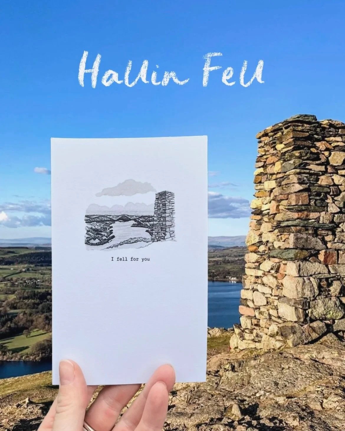 A person holds a white greeting card with a black ink drawing of a landscape on it, with the caption 'I fell for you'. In the background, there is a landscape with a stone structure, a lake, and rolling hills under a clear blue sky. The text 'Halling