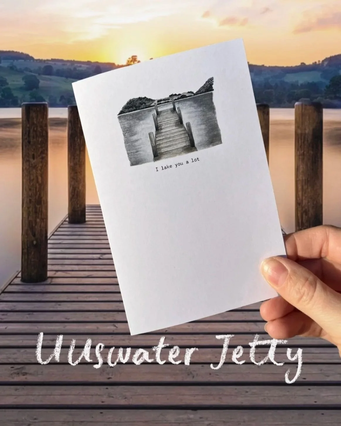 A hand holds a greeting card with a black-and-white drawing of a dock leading to a small island, with the caption "I lake you a lot". In the background, a wooden dock extends over a calm body of water during sunset, with hills and trees in the distan
