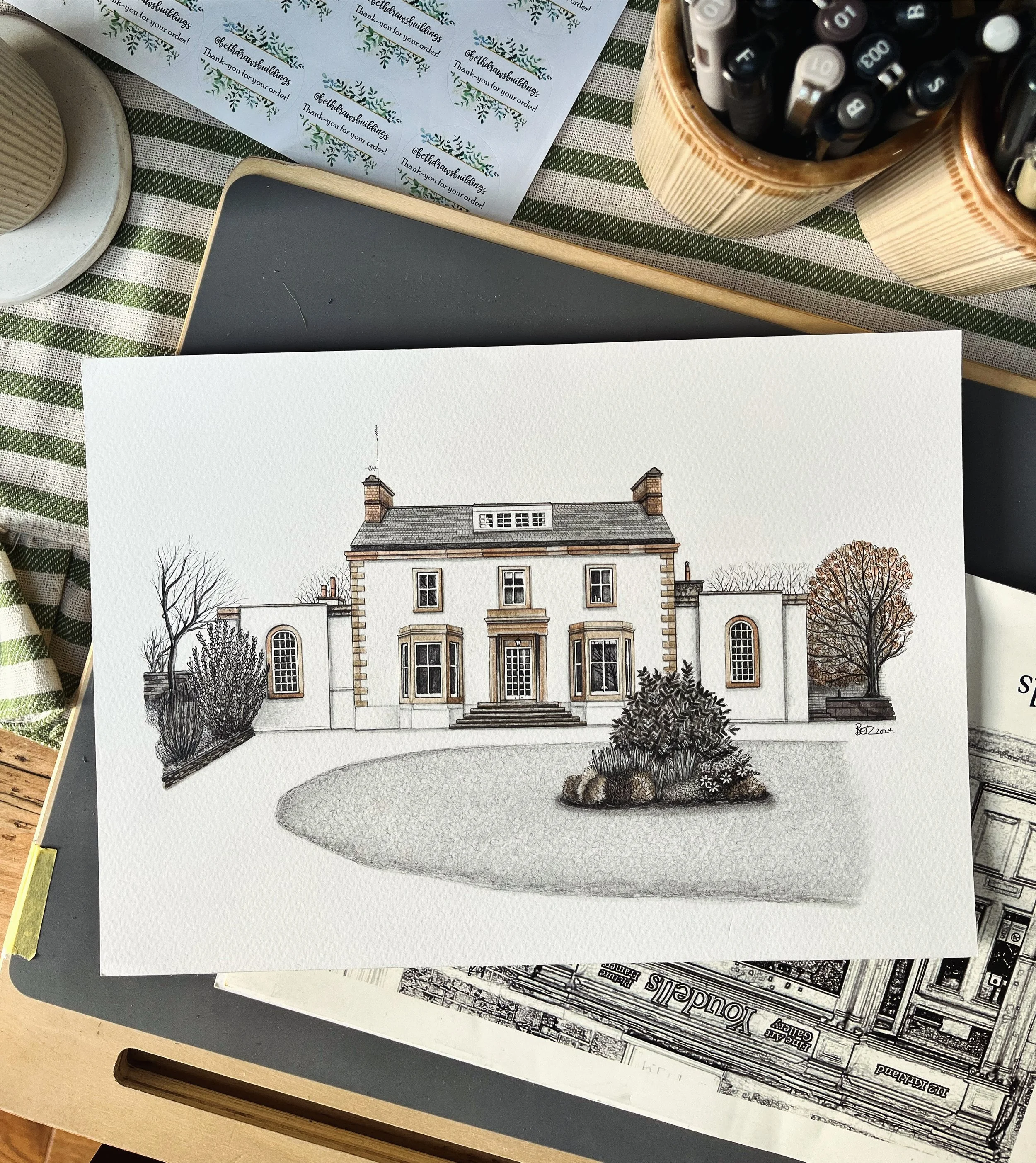 A detailed watercolor illustration of a large, elegant house with a front lawn and trees, set on a table among stationery items and plant-themed thank-you notes.