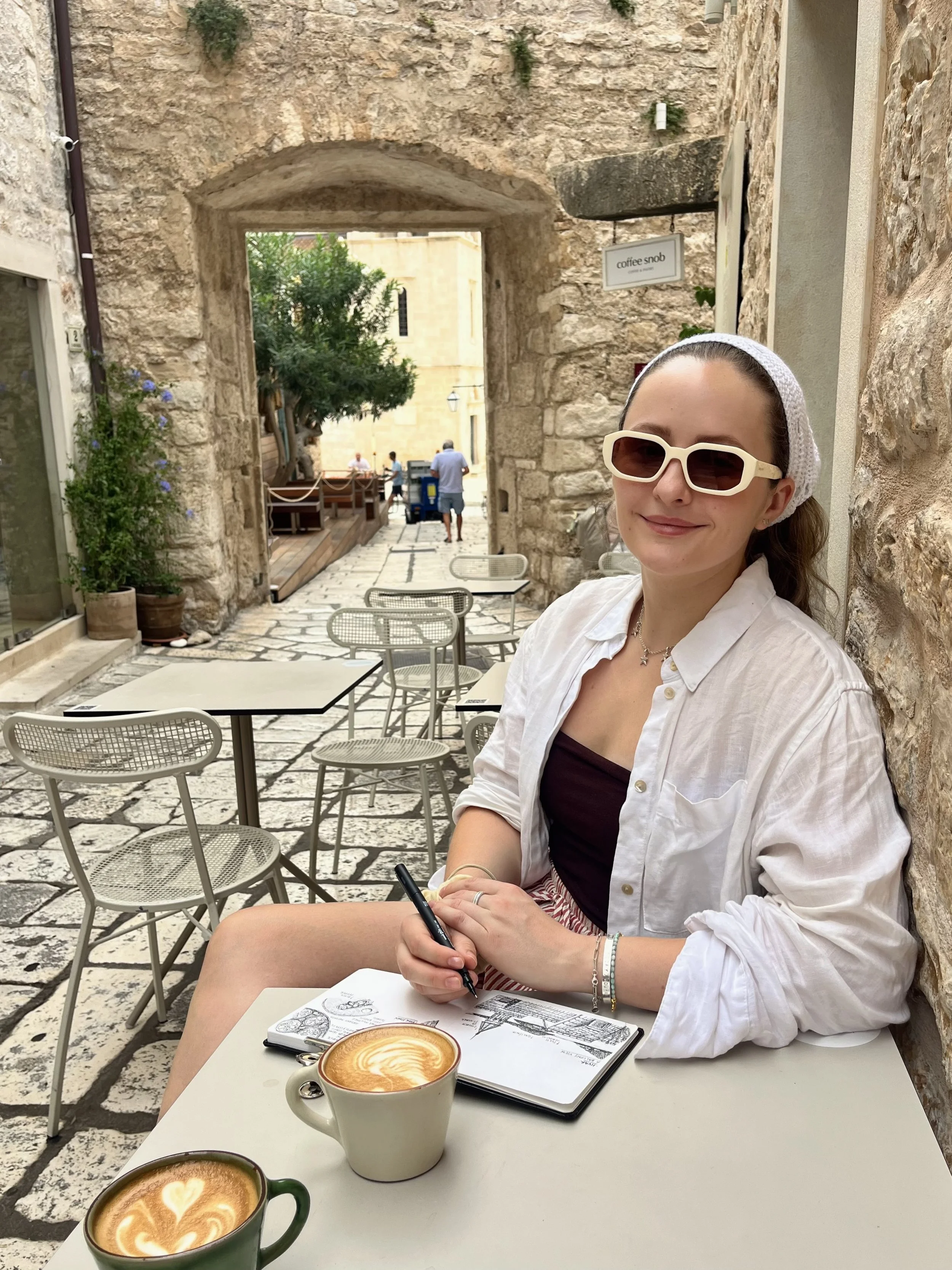 A woman with sunglasses and a white headscarf sitting outside at a café with two cups of coffee, sketching in a notebook, with stone walls and outdoor seating in the background.