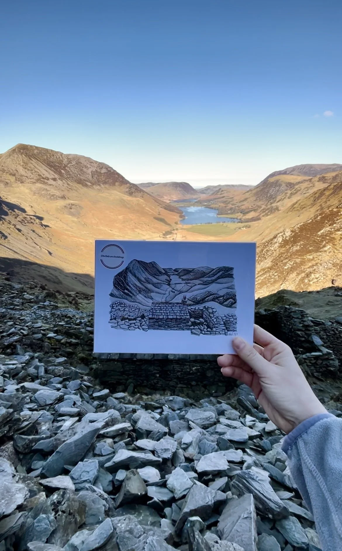 A person holding a sketchbook with a drawing of the landscape behind them, featuring hills, a river, and a lake under a clear blue sky.
