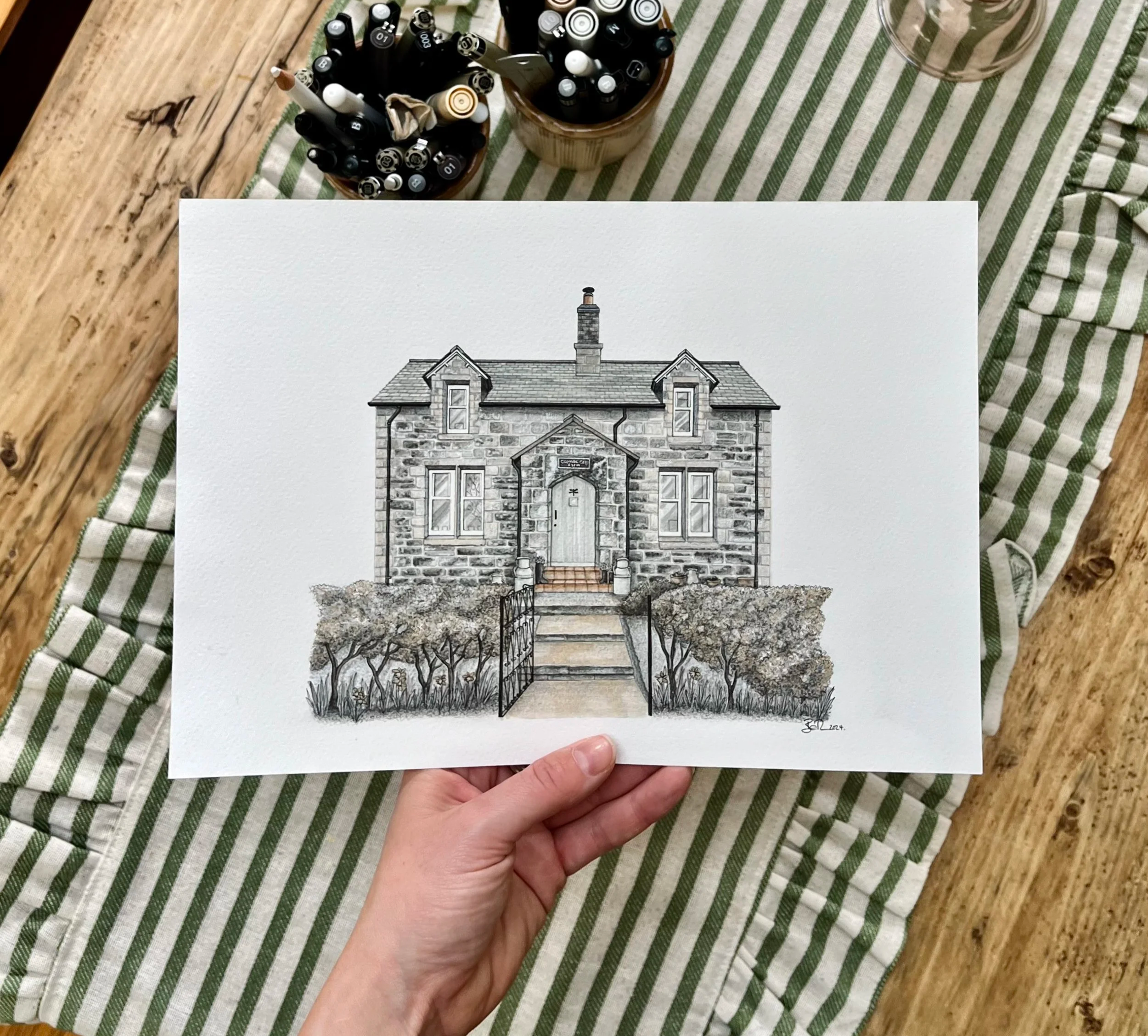 Hand holding a drawing of a stone house with stairs, bushes, and a fence in front, on a striped cloth on a wooden table.