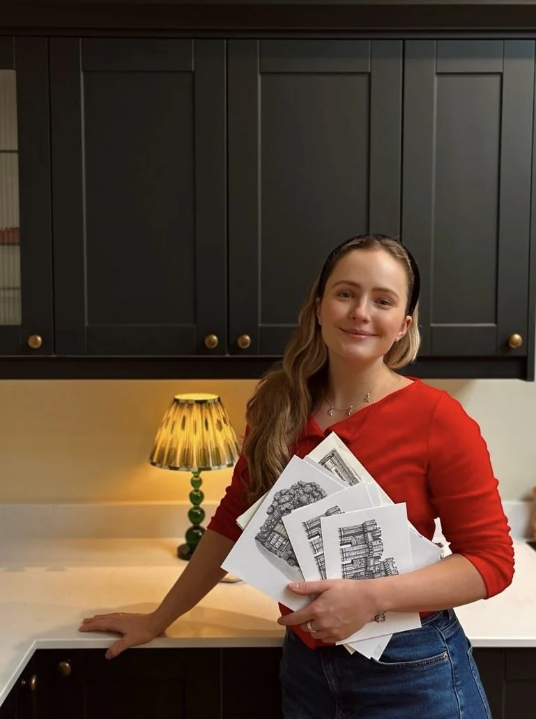 A young woman in a red shirt holding several black and white architectural drawings or plans, standing in a kitchen with black cabinets and a yellow lamp with green base on the counter.