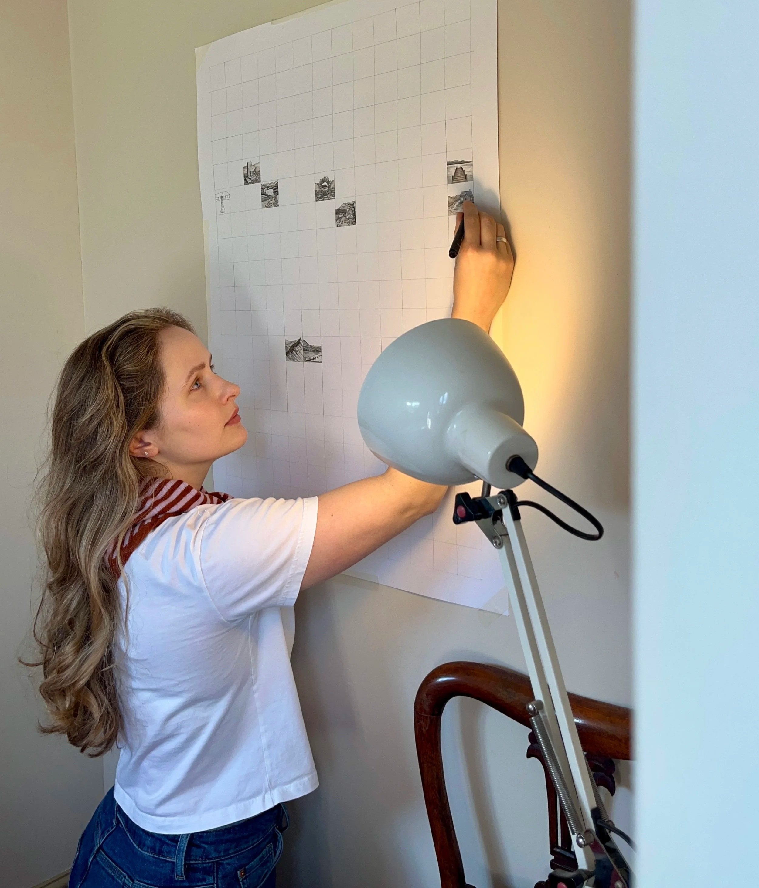 A woman with long wavy hair wearing a white t-shirt with a red and white striped collar and blue jeans is drawing or writing on a large grid paper with black and white images in some of the squares. She is standing next to a white wall, and a lamp on a stand is illuminating her work.