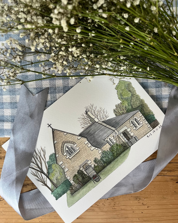 A watercolor illustration of a small stone church with a cross on the roof, surrounded by trees and shrubs, placed on a wooden surface with a checkered cloth and white ribbon, topped with a bunch of white flowers.