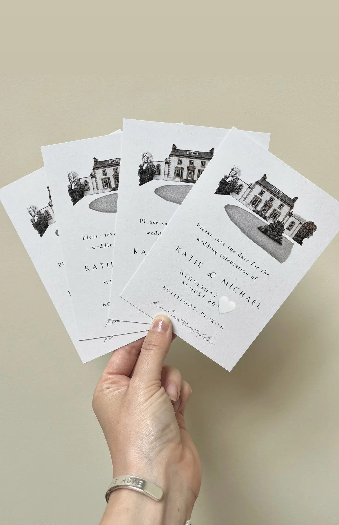 Three wedding save-the-date cards featuring a house illustration, with text including the names Katie and Michael, wedding date August 2023, and location Hollisfoot, Penrith, held by a hand against a plain background.