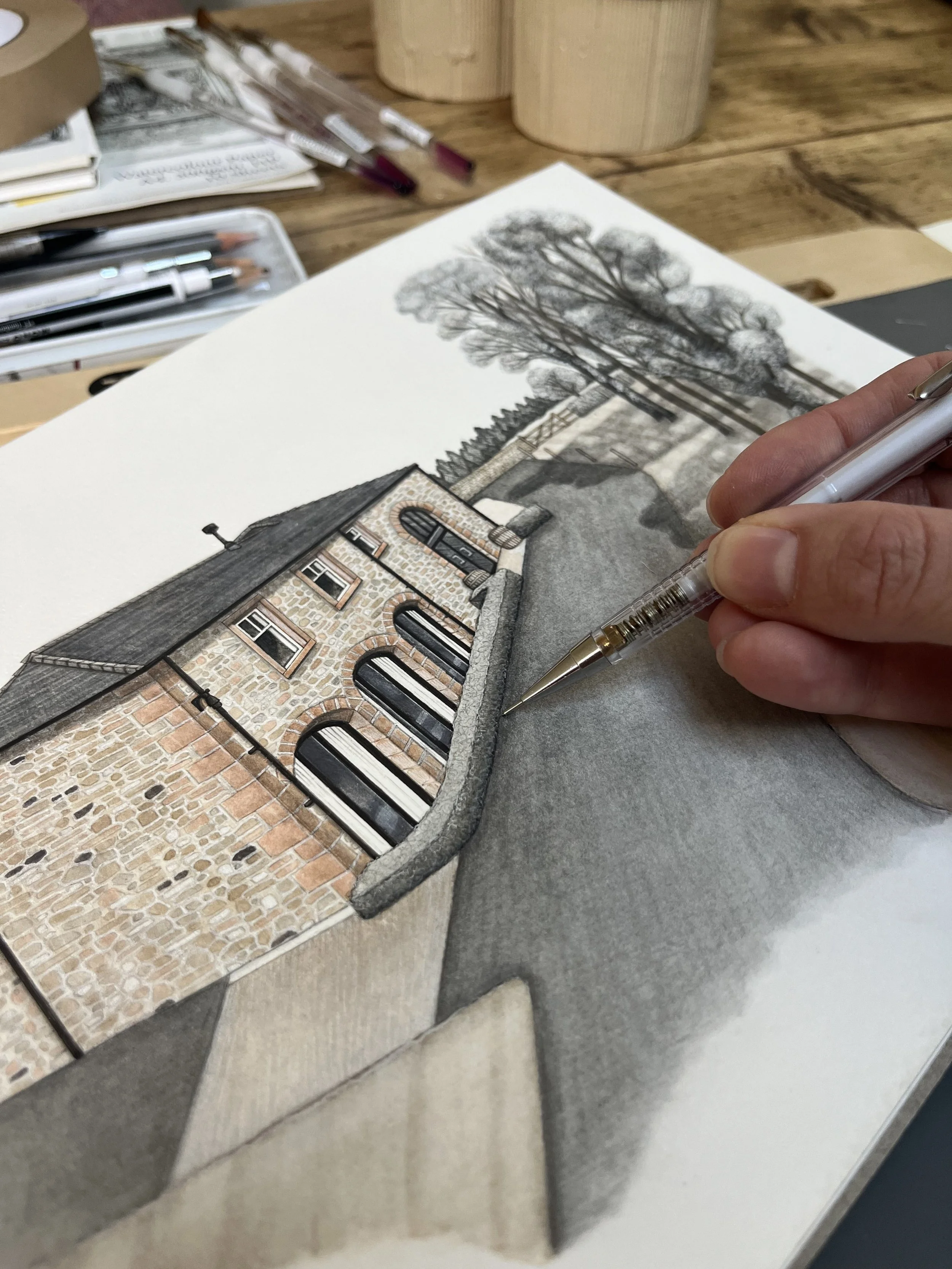 An artist sketches a detailed watercolor painting of a historic brick building with arched windows, surrounded by tall trees, on a wooden desk with art supplies.
