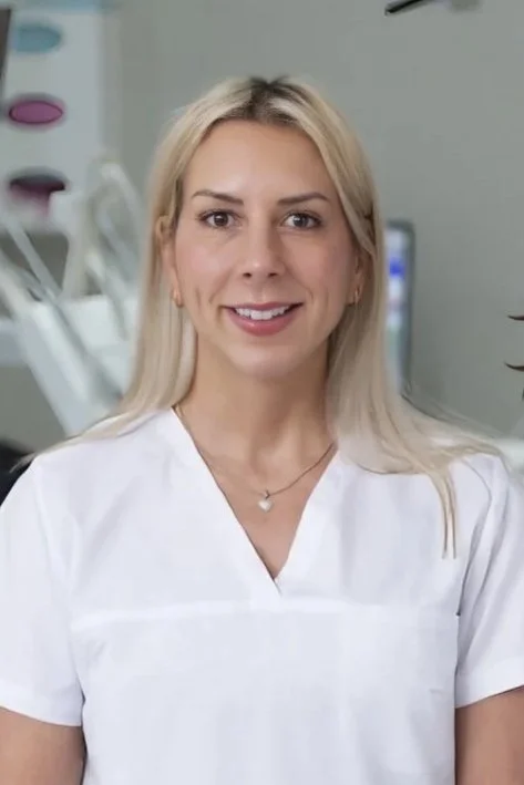 En leende kvinna i vit arbetsuniform står i ett medicinskt miljö med medicinsk utrustning i bakgrunden.