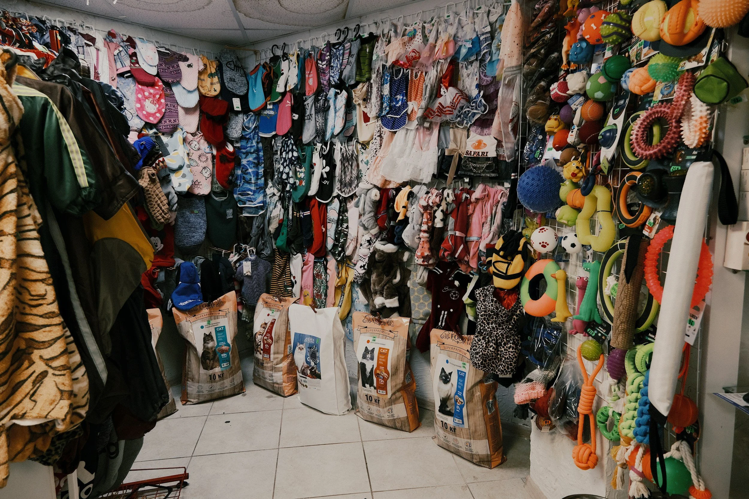 A pet store aisle displaying various pet accessories, toys, and clothing, including bags of cat food, colorful chew toys, and pet apparel.