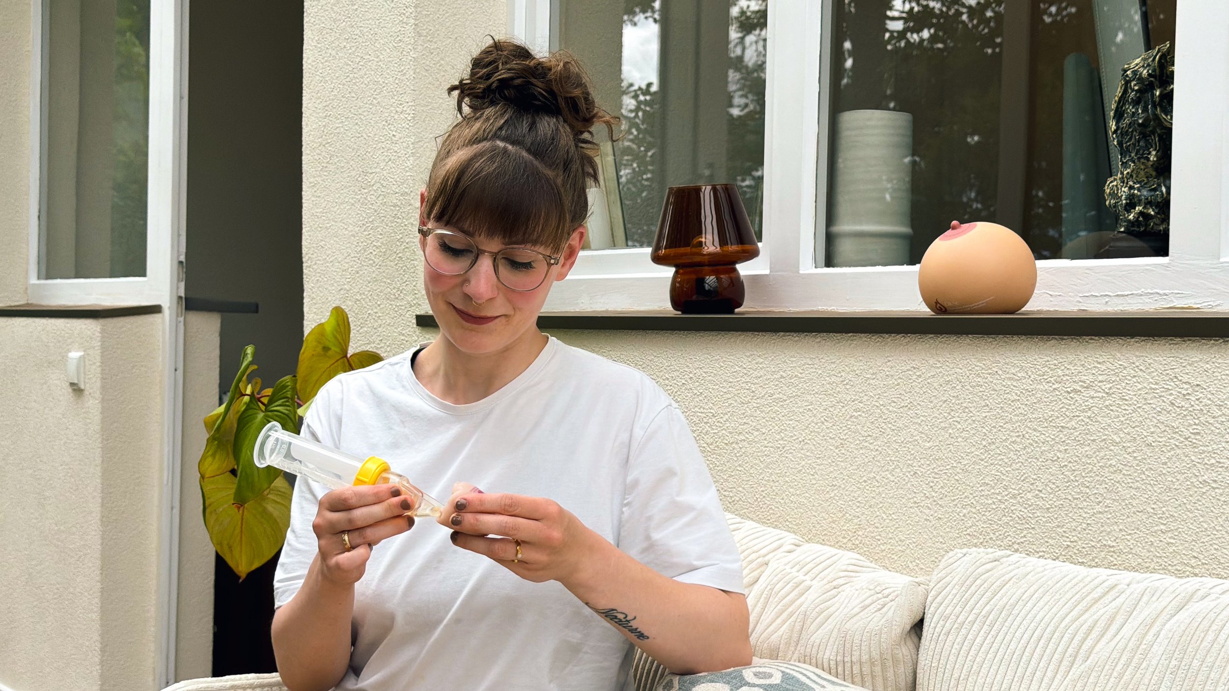 Larissa Brugger alias Lacta Larry, sitzt mit weißem T-Shirt auf einem weißen Sofa und führt mit einem Softcup und einem Silikon Babymundmodel eine stillfreundliche Zufütterung vor.