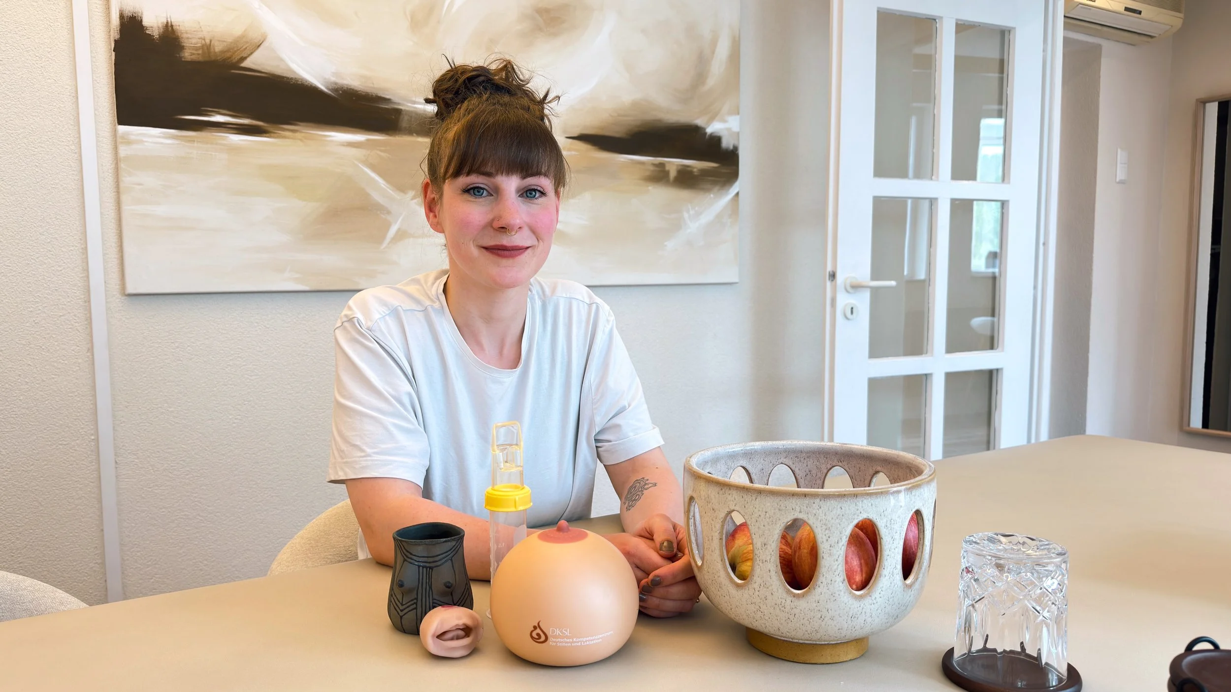 Larissa Brugger alias Lacta Larry sitzt an einem weißen Steintisch, vor ihr eine schöne Keramiktasse mit Brüsten, ein Brustmodell, ein Babymundmodell, ein Softcup und eine Obstschale und im Hintergrund hängt ein Bild.