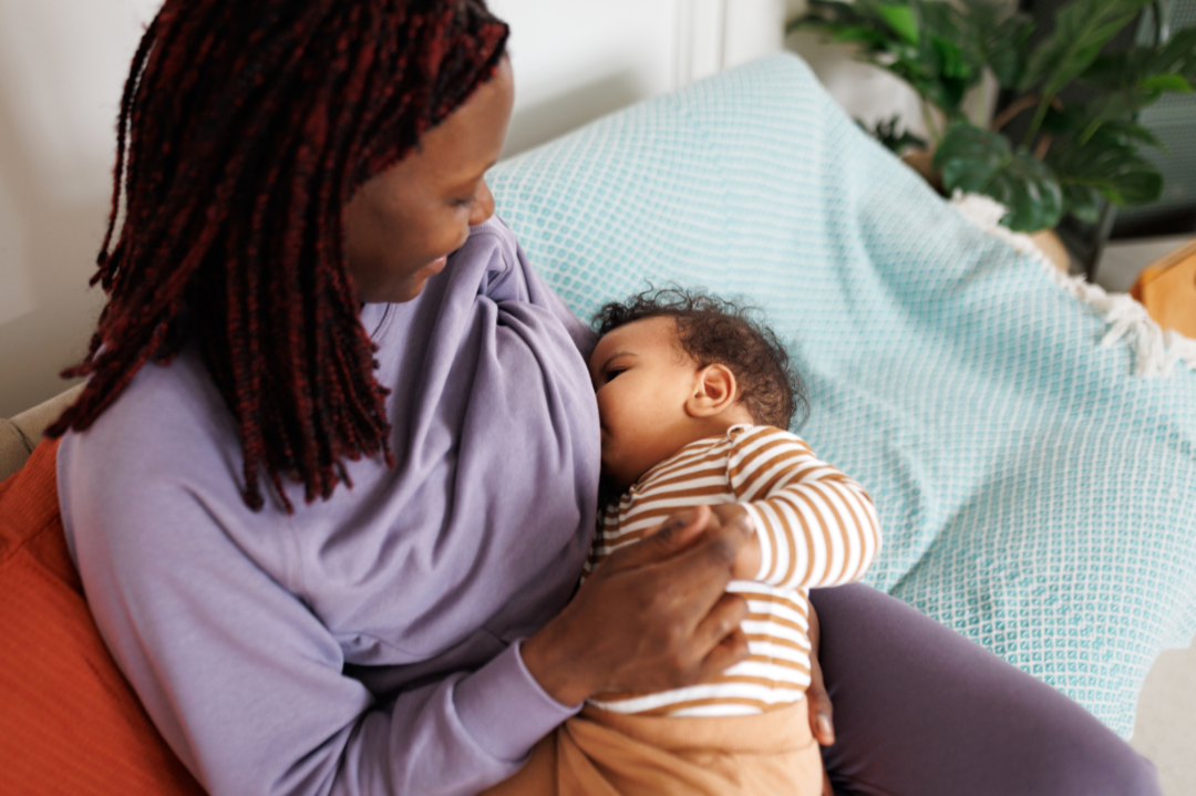 Eine Frau stillt ihr Baby auf dem Sofa, im Wohnzimmer, mit Pflanzen im Hintergrund.