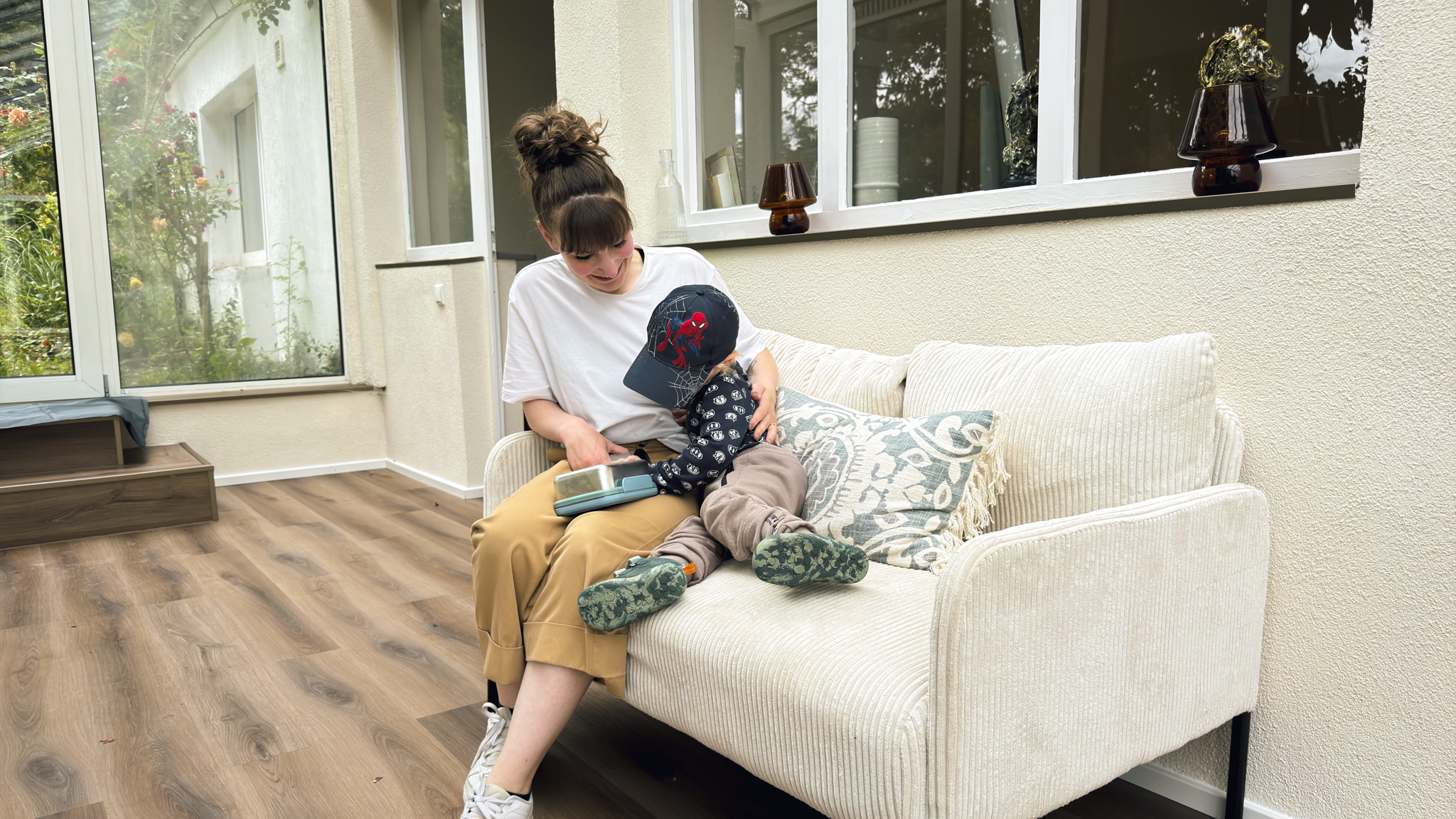 Larissa Brugger alias Lacta Larry sitzt auf einem weißen Sofa, umgeben von ihrem Kind mit Spiderman-Kappe, das teilweise auf ihrem Schoß sitzt.