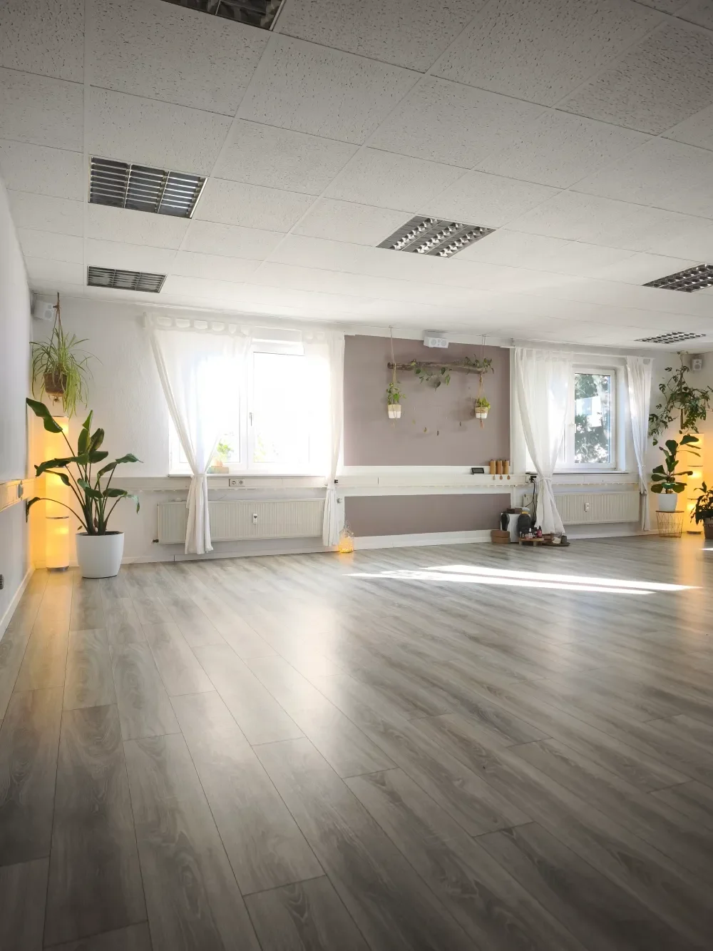 Ein heller, leerer Raum mit Holzfußboden, großen Fenstern mit weißen Vorhängen, Pflanzen und sanfter Beleuchtung. Ein Yoga und Kursraum des Mind&Body Ateliers in Ulm in dem das Stillcafé vor Ort stattfindet.