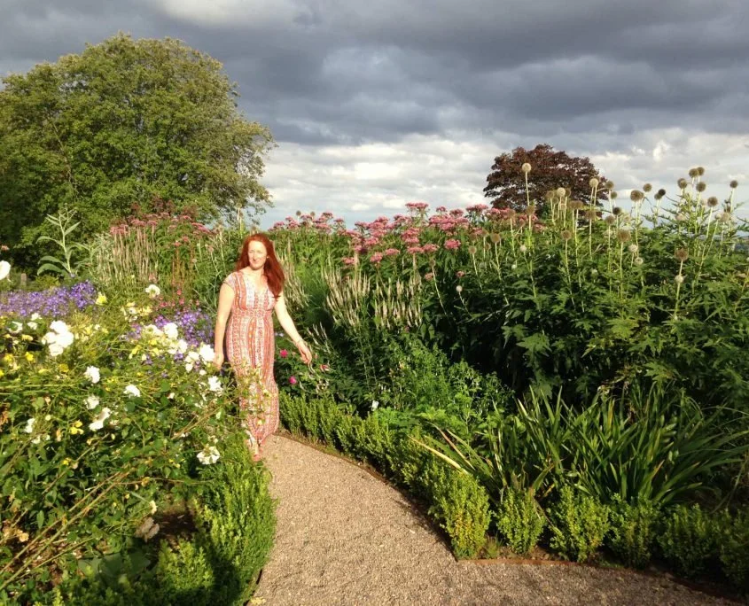 A woman in a long dress walking down a path towards the camera, flanked by tall flower borders edged with hedging.