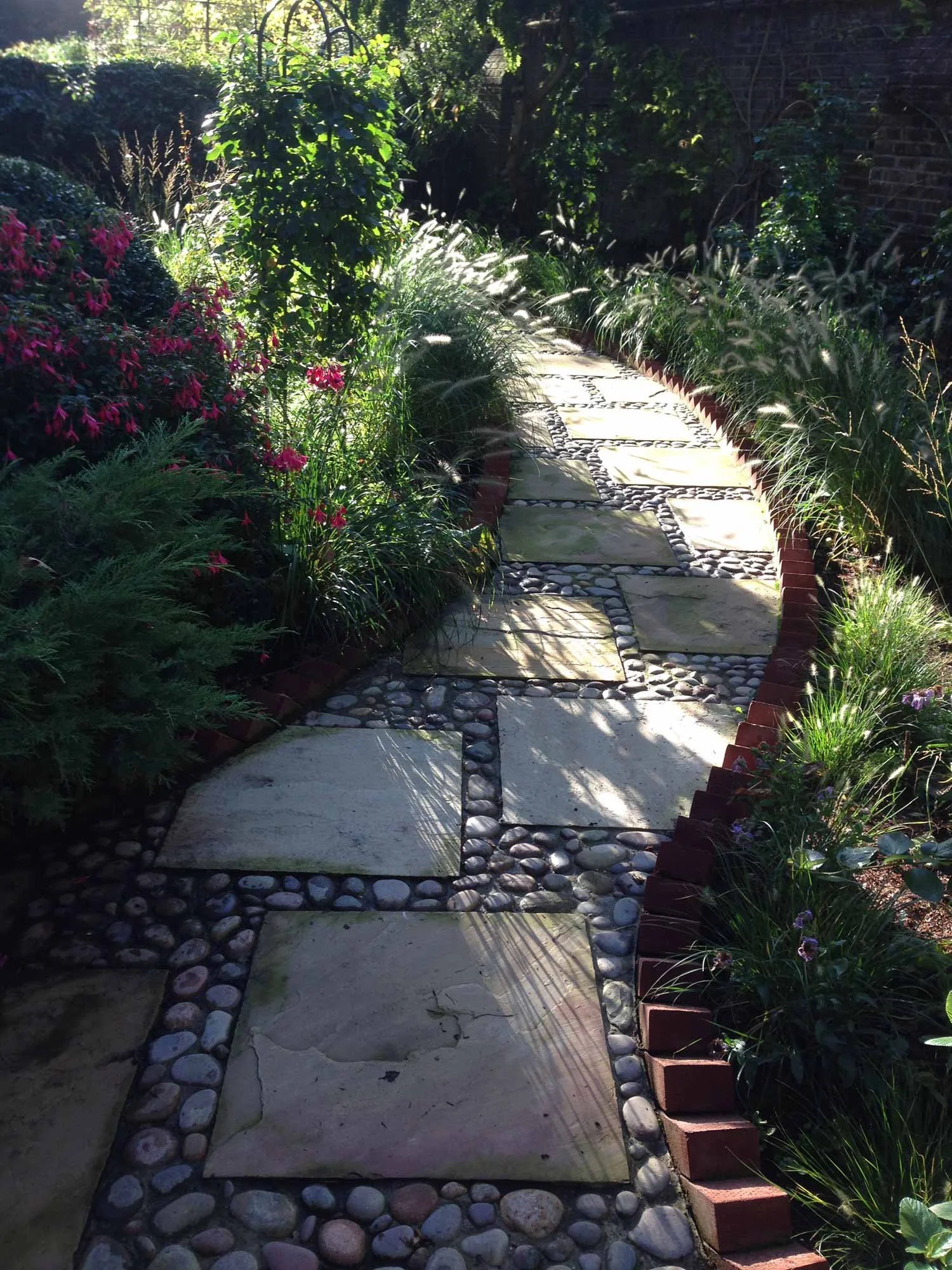 A winding path with cobbles, flagstones and terracotta edging, flanked by colourful borders, at a garden at Chelwood House, London.