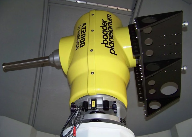 A yellow Hubble Phenoid telescope component with the words 'NASA DOD' and 'Hubble Phenoid' printed on it, attached to a black mounting bracket with circular patterns, located indoors.