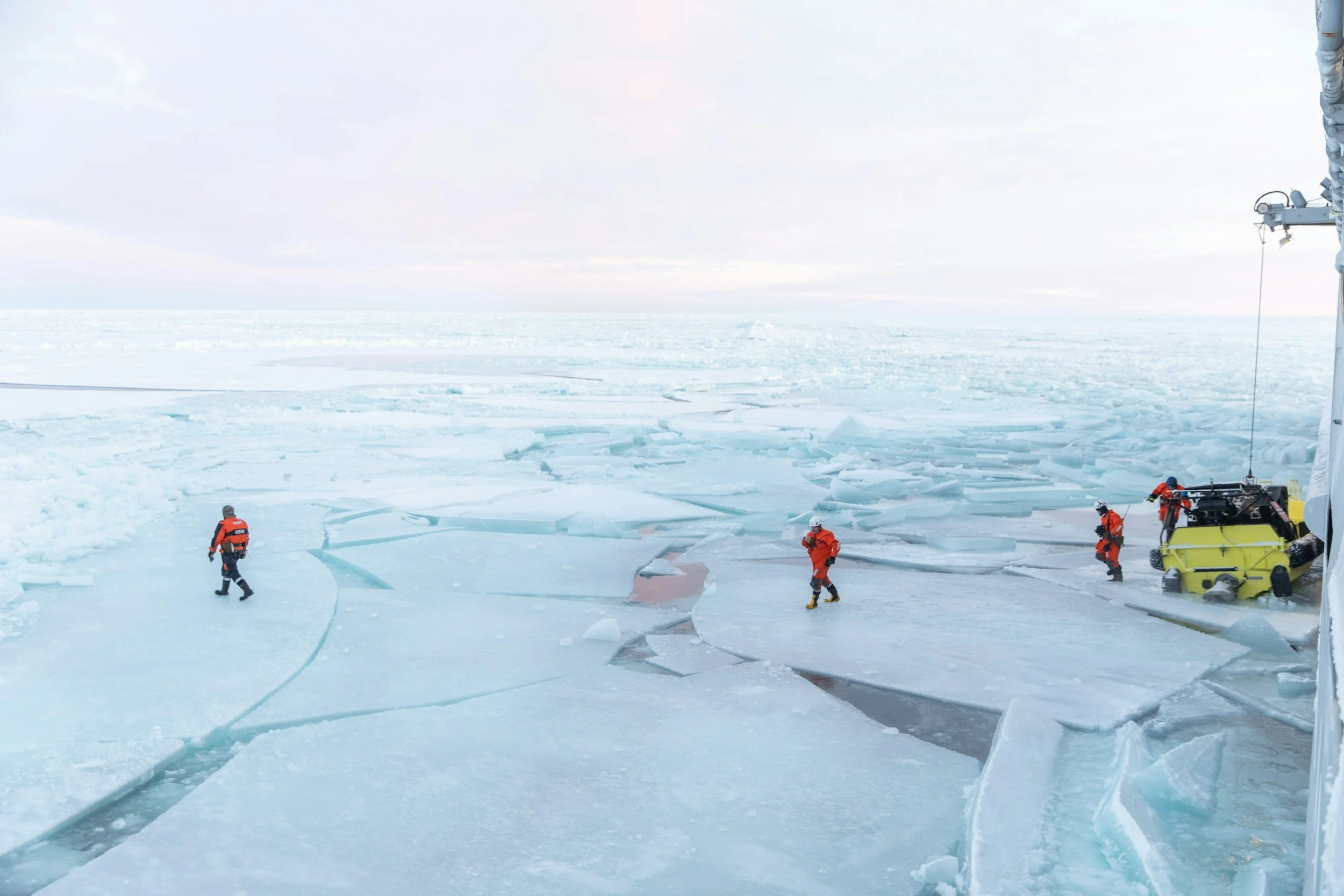 Scientists or researchers in orange and black cold-weather clothing working on ice-covered terrain, with large cracks and broken ice sheets, near a crane and equipment.