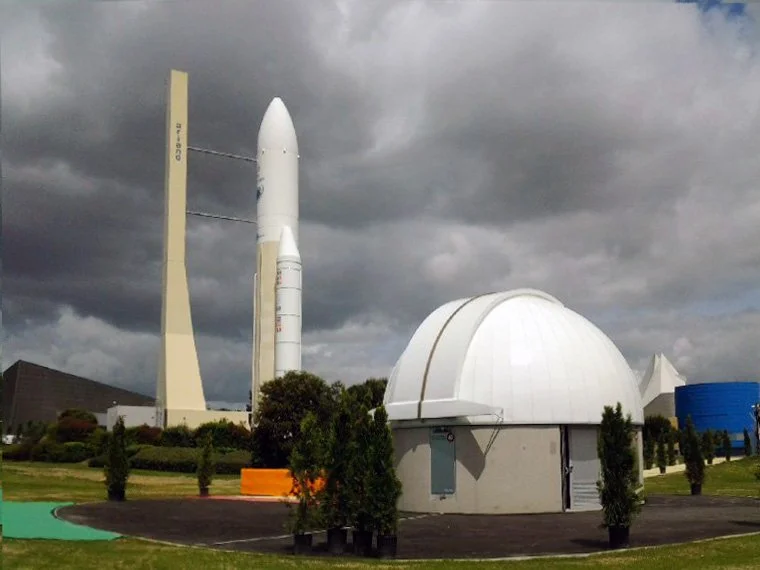 A space launch facility with a tall rocket on a launch pad and an observatory dome in the foreground, under a cloudy sky.
