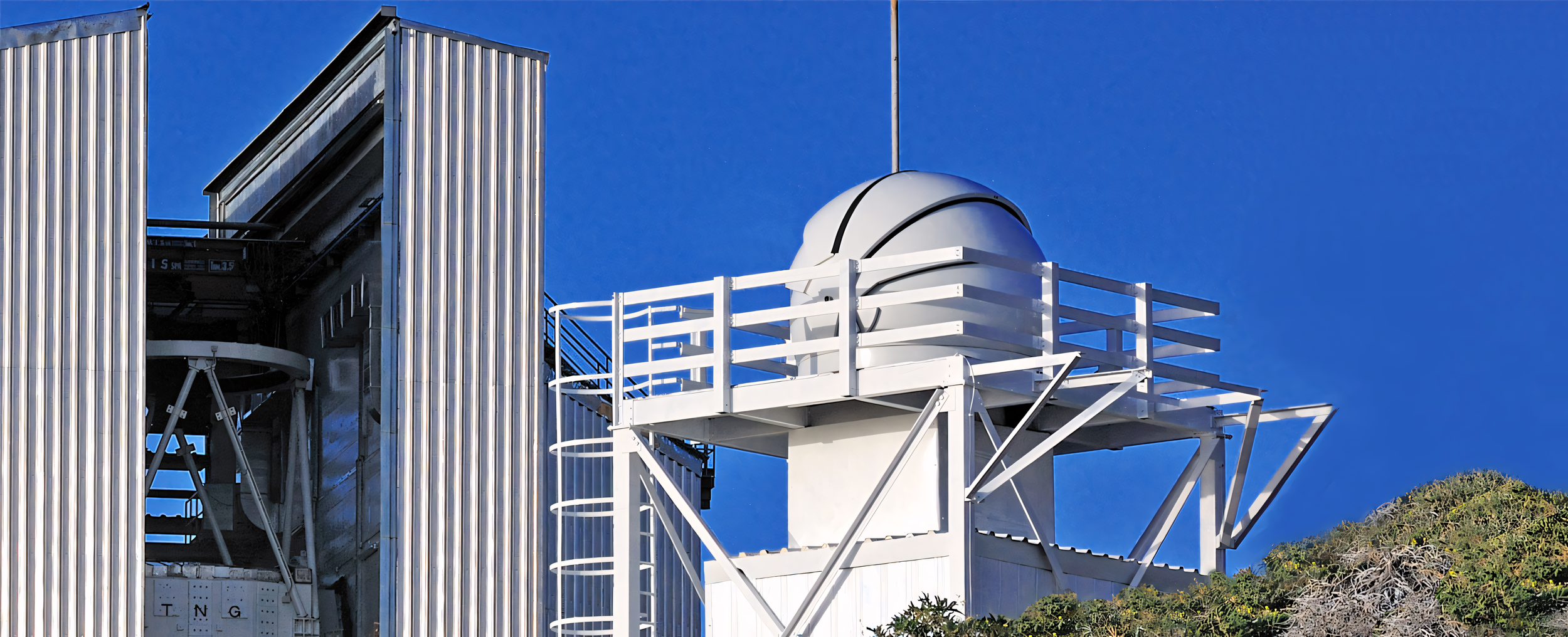 A large telescope with a white dome and metal support structure, set against a clear blue sky, surrounded by greenery.