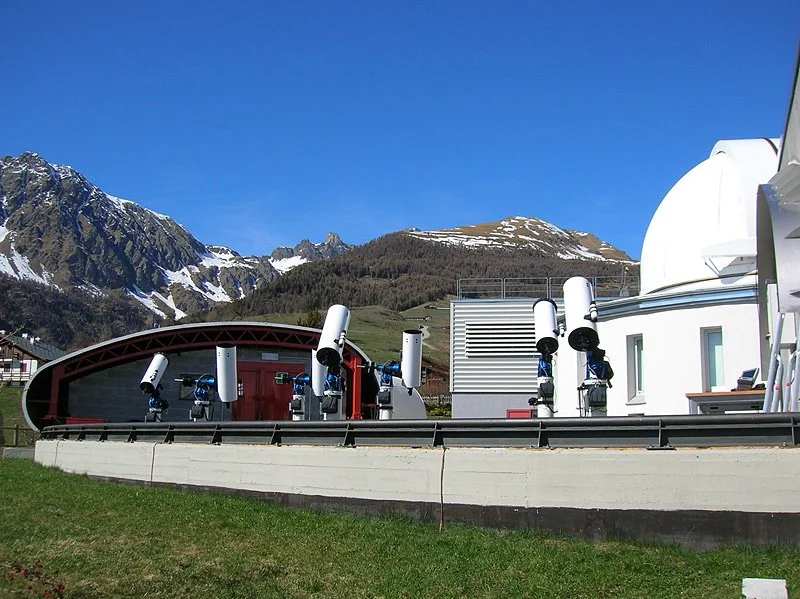 Astronomical observatory with multiple telescopes on a rooftop, mountain landscape in background under clear blue sky.