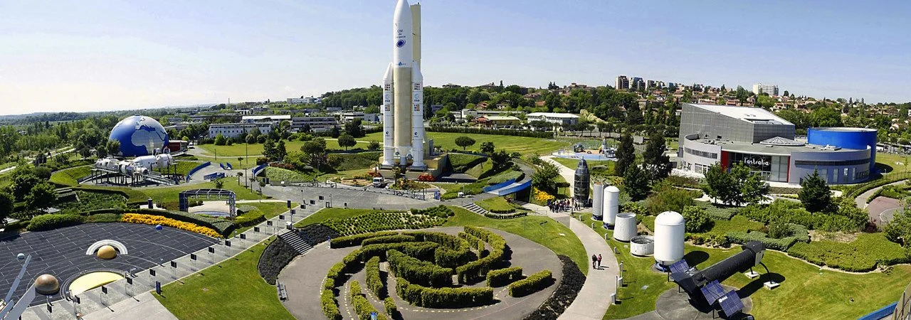 A large science and space museum with a rocket and planetarium, featuring a landscaped outdoor area with pathways, bushes, and scientific exhibits.