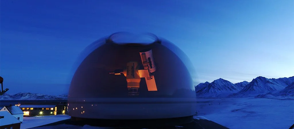 A large observatory dome on snowy mountains under a twilight sky reflecting a telescope inside.