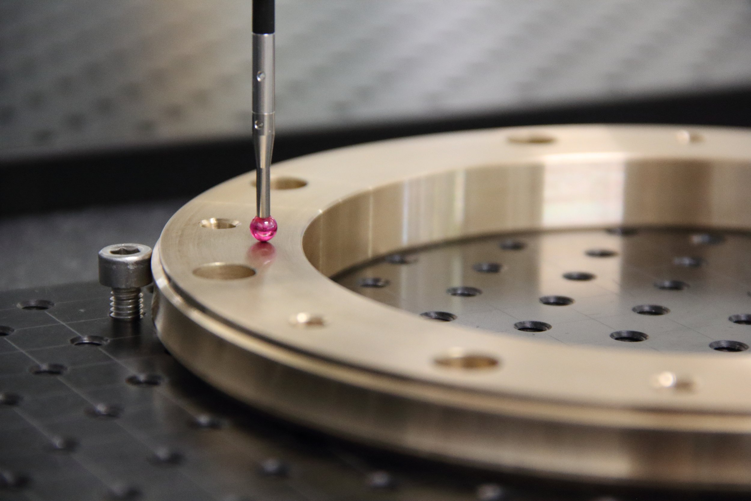 Close-up of a laboratory or industrial machinery with a metallic circular component and a pipette depositing pink liquid into a small drop. The machinery has a matte black surface with multiple holes and a metallic mounting screw.