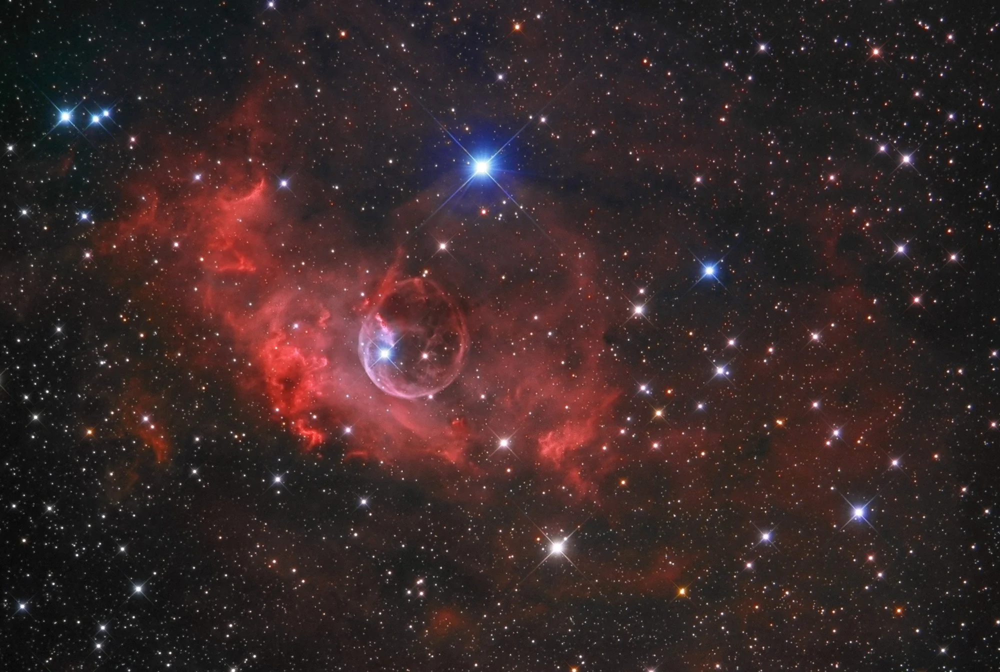A deep space image showing numerous stars, a red nebula, and a bright blue star near the center.