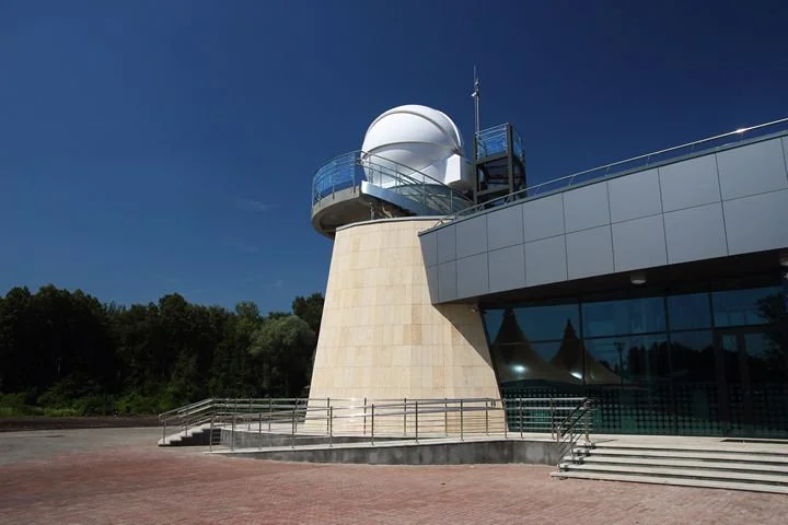 A modern observatory with a white dome and viewing deck, connected to a building with large glass windows, set against a clear blue sky.