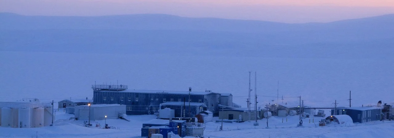 A remote snowy research station with multiple small buildings and antennas at dusk or dawn, surrounded by snow and ice.