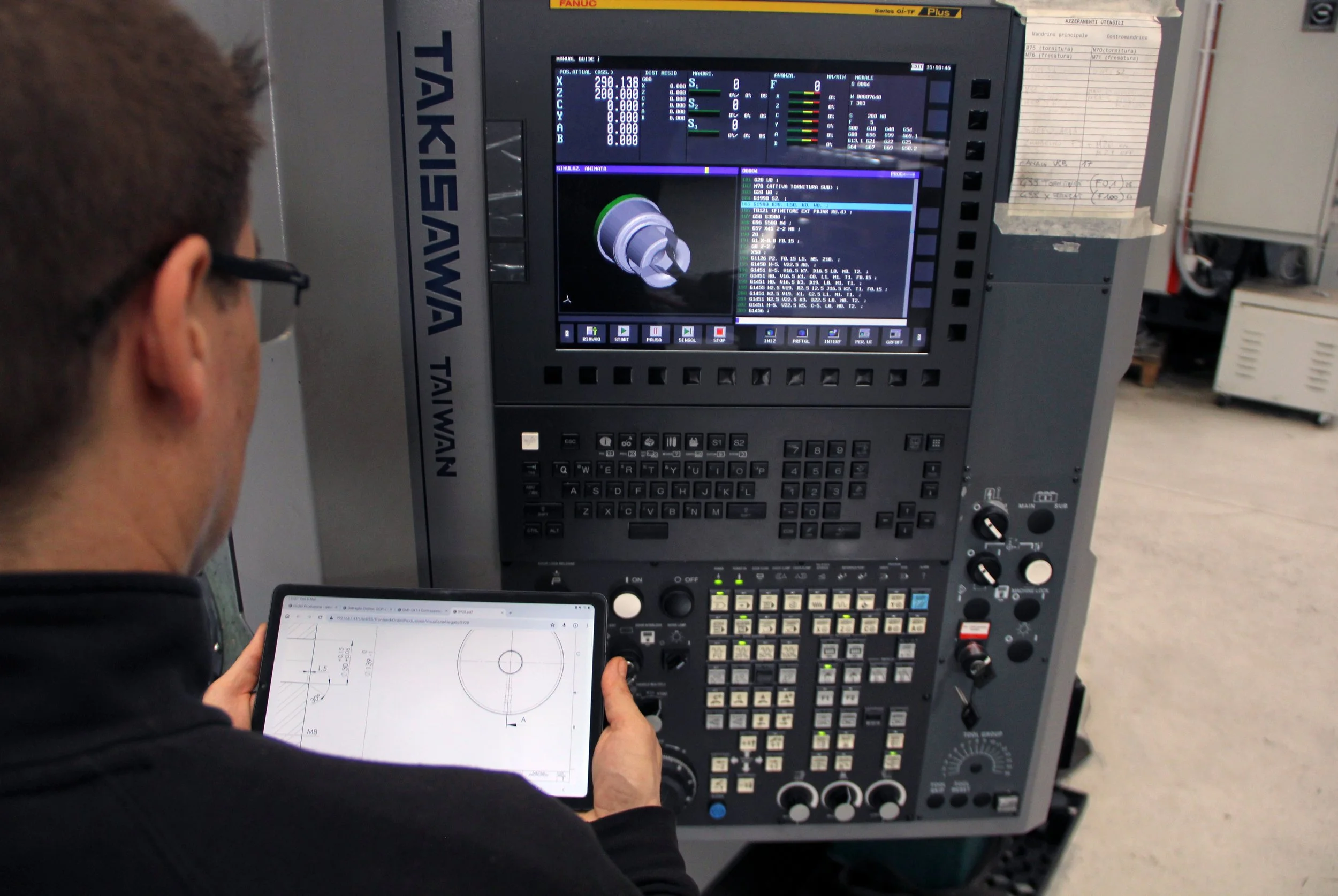 A person operating a CNC machine, looking at a tablet with technical drawings and the machine's control panel with a display showing a 3D model of a part.