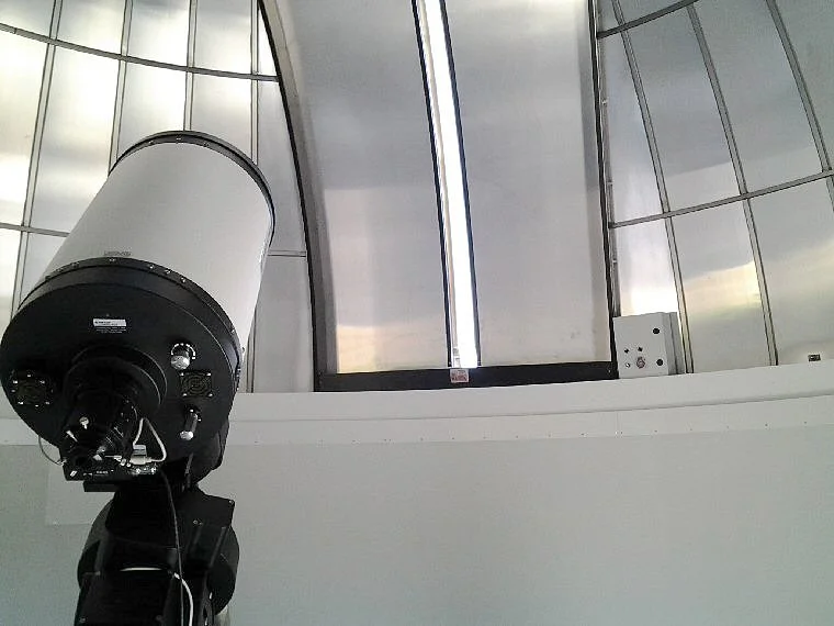 An astronomical telescope mounted in an observatory with a glass ceiling and walls, allowing natural light to enter.