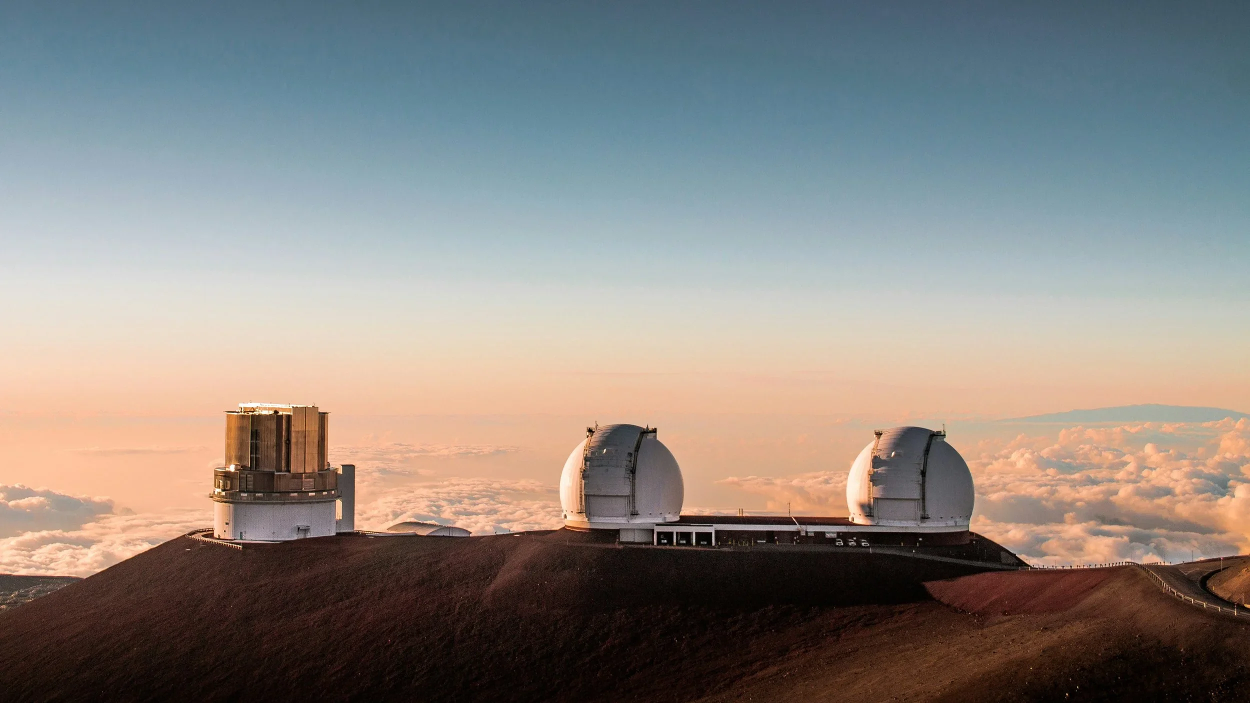 High-altitude view of observatory telescopes on a mountain, with clouds and a sunset sky in the background.