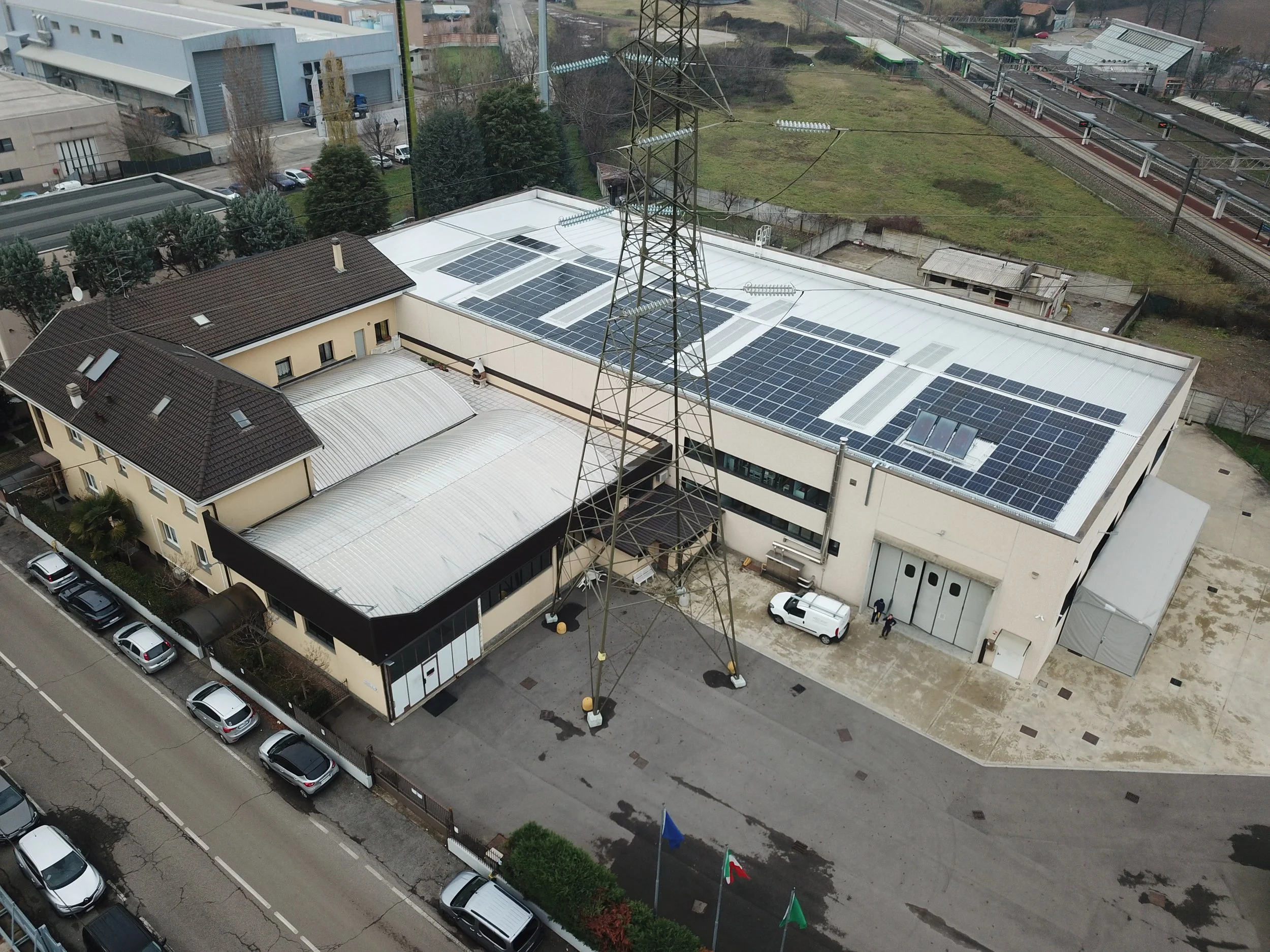 Aerial view of COMEC Company (10Micron) building with solar panels on its flat roof, an adjacent building with a curved roof and a parking lot. Residential and industrial buildings, train tracks, and greenery are in the background.
