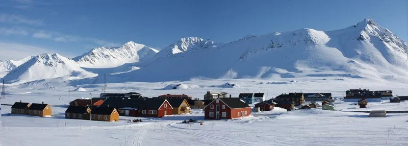 A small village with colorful houses in a snowy landscape, with snow-covered mountains in the background under a clear blue sky.