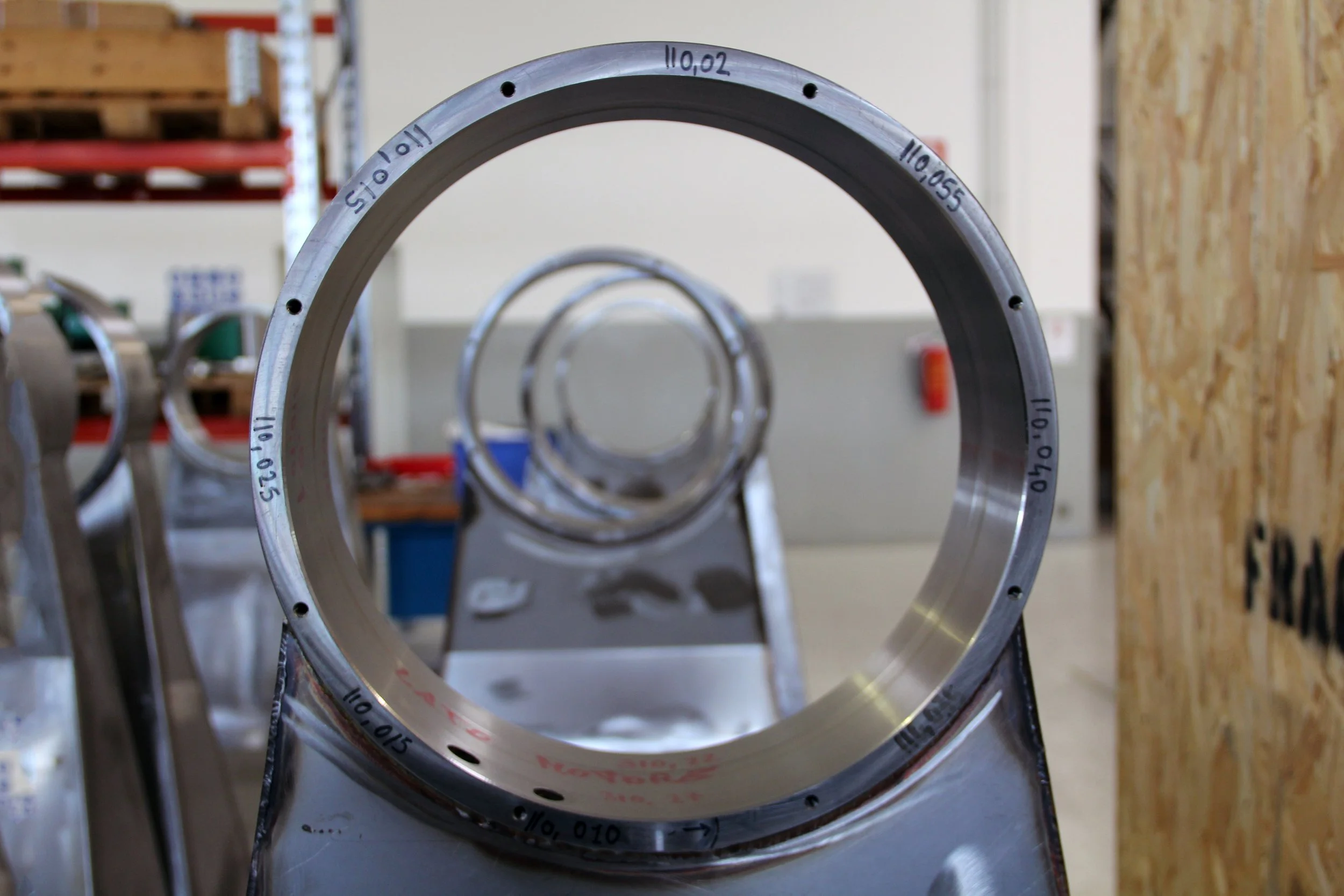 Close-up of a metal circular piece with numbered markings, placed on a stand in an industrial or warehouse setting, with additional similar pieces in the background.
