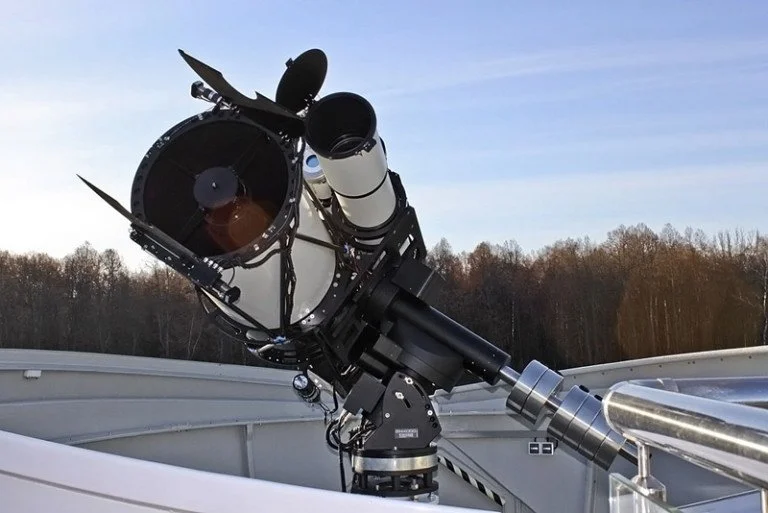 Large telescope mounted on an observatory rooftop with a clear sky and trees in the background.