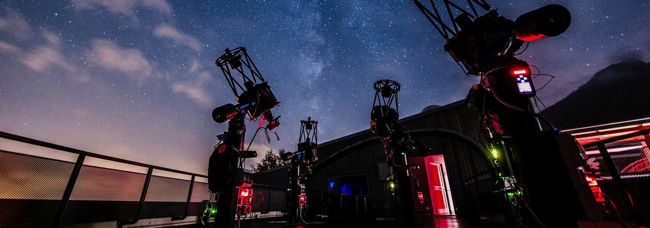Night sky with stars and Milky Way, telescopes set up on a rooftop observatory with a railing, building with illuminated door, and mountain silhouette in the background.