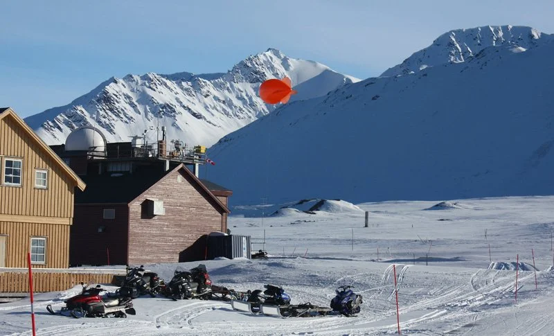 Snow-covered alpine landscape with wooden buildings, parked snowmobiles, and mountains in the background, with an orange parachute floating in the sky.