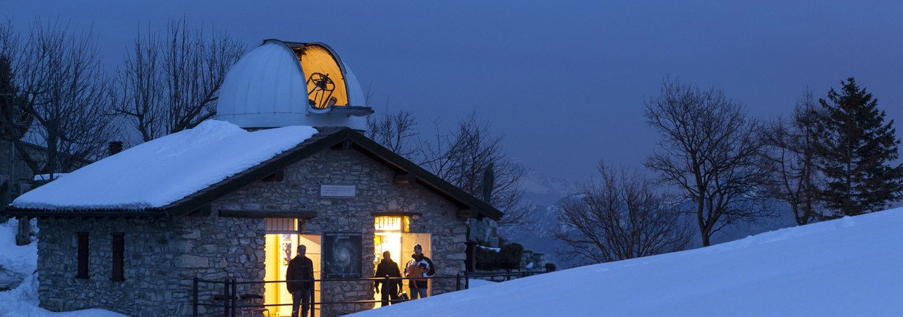 A snowy landscape with a small stone observatory building at dusk, people are standing outside with lights on inside.
